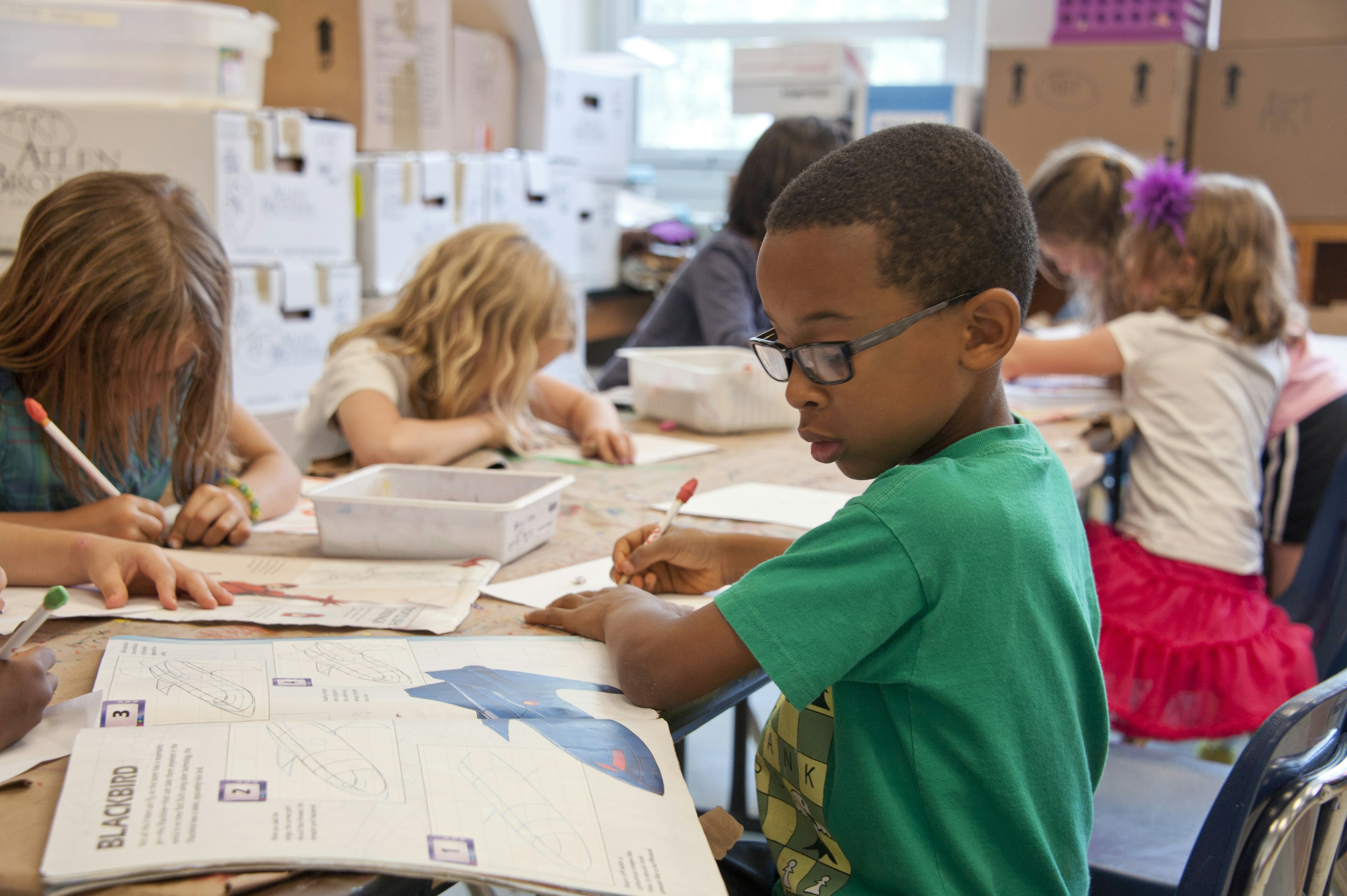 A boy in glasses works carefully through a drawing workbook at an art table